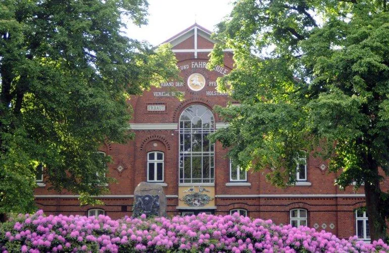 Historische Backsteinfassade der Reit- und Fahrschule des Holsteiner Verbandes in Elmshorn mit Giebelinschrift, Holsteiner Pferdewappen, großem Rundbogenfenster und blühenden Rhododendren davor